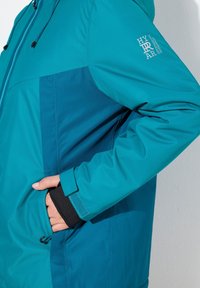 Tealfärgad och mörkblå väderbeständig jacka med justerbar huva, sidofickor, elastiska ärmslut och logotypdetalj på ärmen.