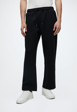 Homme portant un pantalon de survêtement noir avec cordon de serrage et des baskets blanches, debout devant un fond clair uni.