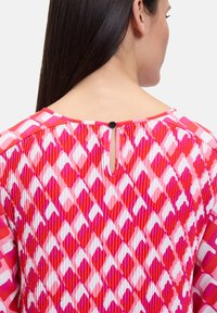 Blusa colorata con motivo a diamante in tonalità di rosso, rosa e bianco. Presenta un dettaglio a chiave sul retro con chiusura a bottone tondo.