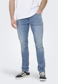 Jeans in denim azzurro chiaro con una vestibilità slim, dettagli di scolorimento sottili, design a cinque tasche, abbinati a sneakers nere con strisce bianche.