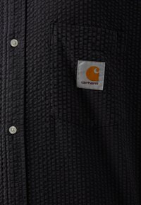 Camicia nera button-up con un motivo a griglia testurizzato, una tasca sul petto con un'etichetta bianca e un accentuazione arancione prominente.
