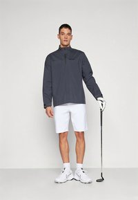 Giacca grigia con mezza cerniera e dettagli neri, pantaloncini bianchi e mazza da golf. Modello con scarpe da ginnastica bianche e guanto da golf, in piedi su sfondo neutro.