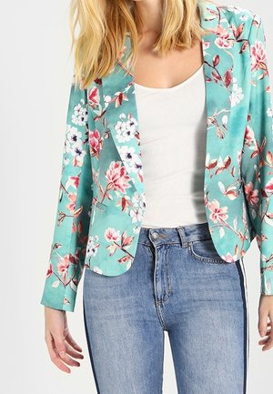 Kvinde iført en turkis blomsterblazer over en hvid top og blå jeans med sidelister mod en ensfarvet baggrund.