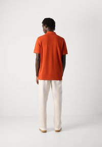Mann som står med ryggen til, iført en kortermet oransje polo shirt, hvite bukser og hvite sko med lysebrune såler, foran en enkel hvit bakgrunn.