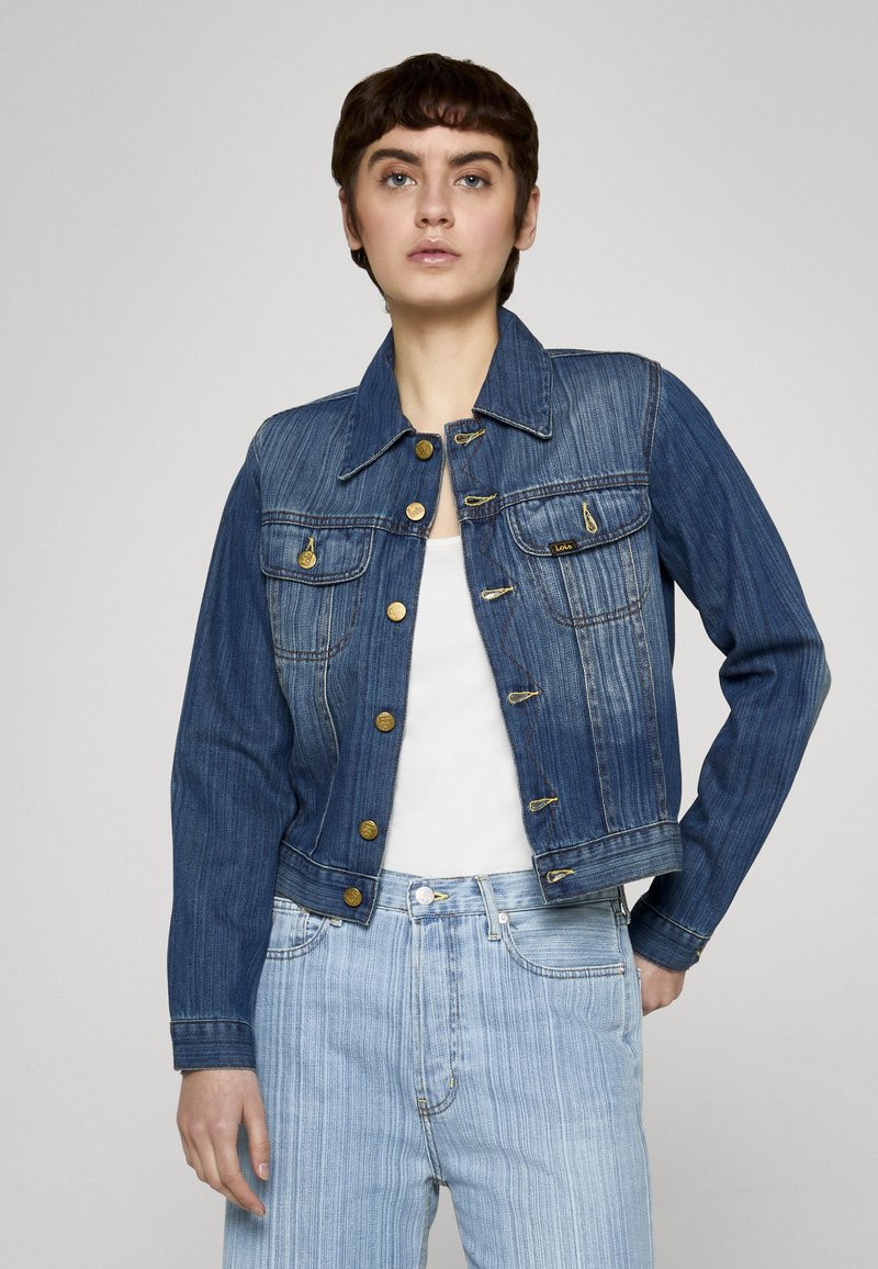 Riflová bunda tmavomodrej farby s klasickým golierom, štyrmi prednými vreckami, zlatou kovovou výbavou a skráteným strihom, skombinovaná so svetlomodrými džínsami.