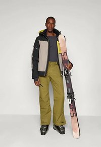 Χειμερινά ρούχα για σκι με πολύχρωμη τζάκετ σε μαύρο, γκρι και κίτρινο, συνδυασμένα με ελαιόχρωμο παντελόνι και μαύρες μπότες σκι, κρατώντας σκι.