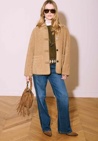 Veste en polaire couleur fauve avec boutons, pull vert en dessous, jean évasé bleu, et chaussures pointues marron. Tenant un sac à franges marron.