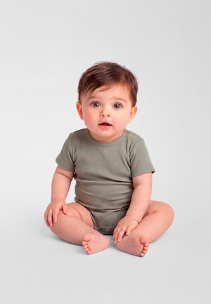 Småbarn med brunt hår og grå heldrakt som sitter med korslagte ben på en ensfarget lys grå bakgrunn, ser rett frem med munnen lett åpen.