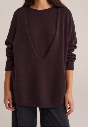 Vrouw draagt een donkerpaars oversized gebreid mouwloos vest met V-hals over een bijpassende trui met lange mouwen en zwarte broek.