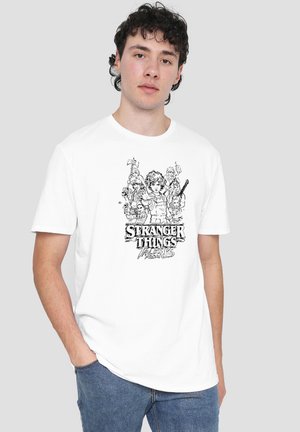 Young man wearing white t-shirt with black Stranger Things graphic and characters, standing with one hand in pocket against gray background.