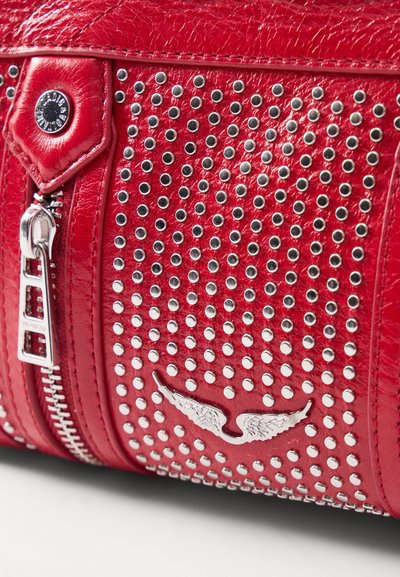 Sac en cuir rouge avec des clous en argent, doté d'une fermeture éclair et d'un emblème décoratif en forme d'aile. Surface texturée et accents en métal brillant.
