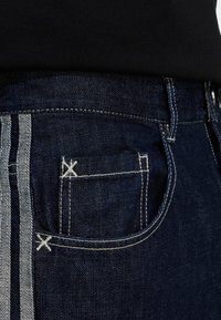 Temno modre jeans hlače iz jeansa s kontrastnimi svetlo modrimi stranskimi paneli, poudarjenimi šivi in majhnim, podrobnim zadnjim žepom.