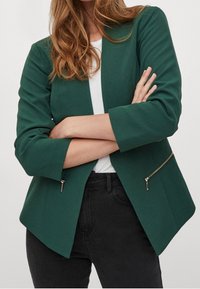 Grön kavaj med skräddarsydd design, med spetsig nederkant, trekvartslånga ärmar och fickor med dragkedja på sidorna. Slät tygtextur.