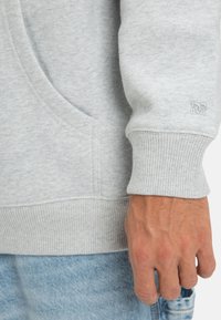 Sudadera gris clara con un bolsillo canguro, puños acanalados y dobladillo inferior. Presenta una textura sutil con una etiqueta de logo en la manga.