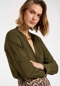 Blusa verde oliva con scollo a V e maniche lunghe, abbinata a una gonna a fantasia con motivi geometrici in marrone, oro e crema. Collana d'oro.