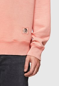 Light pink sweatshirt featuring ribbed cuffs, a textured fabric, and a small label with a graphic design on the front lower left.