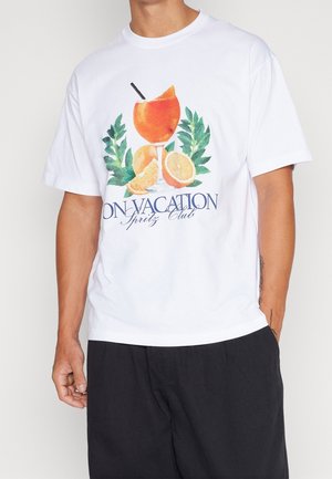 T-shirt en coton blanc avec un graphique coloré représentant un verre à cocktail, des tranches d'orange et des feuilles vertes. Le texte indique "EN VACANCES Spritz Club."