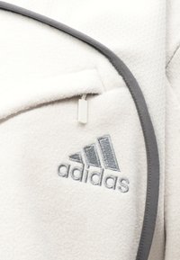 Λευκό φλις ύφασμα με γκρι λεπτομέρειες, που διαθέτει κλειστό τσέπη με φερμουάρ και κεντημένο λογότυπο Adidas σε γκρι. Είναι ορατό υλικό με υφή και εξαγωνικά σχέδια.