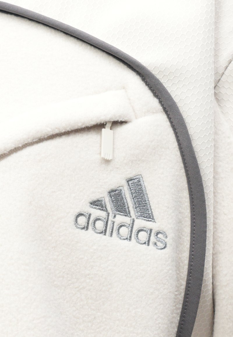 Witte fleece stof met grijze accenten, met een ritszak en een geborduurd Adidas-logo in grijs. Textuur in hexagonale vorm is zichtbaar.
