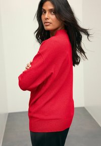 Pull rouge, en maille douce, coupe décontractée, épaules tombantes, ourlet et poignets côtelés. Porté avec un pantalon noir, vu de côté.
