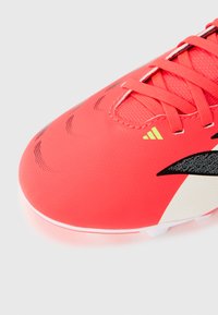 Φωτεινό κόκκινο ποδοσφαιρικό παπούτσι με μαύρες και λευκές λεπτομέρειες σχεδίου, κόκκινες κορδόνια και ένα μικρό κίτρινο λογότυπο κοντά στη γλώσσα, σε λευκό φόντο.