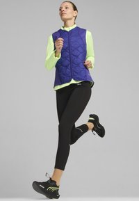 Purpurinis siuvinėtas liemenė virš neoninės žalios ilgomis rankovėmis palaidinės, derinama su juodomis leginsais ir juodais batais su neoniniais akcentais, esanti judesyje.