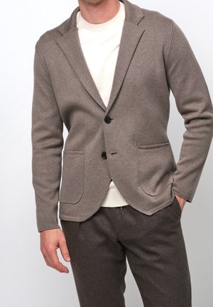 Mężczyzna w taupe'owym dzianinowym blezerze z czarnymi guzikami, białej koszulce z okrągłym dekoltem i ciemnotaupe'owych spodniach, z jedną ręką w kieszeni, na jednolitym tle.