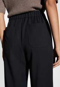 Pantaloni neri con cintura elastica e due tasche posteriori. Il tessuto è liscio con una leggera lucentezza, caratterizzato da una vestibilità ampia e rilassata.