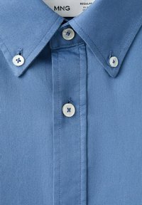 Blauw overhemd met knopen en een puntige kraag, met een close-up van de textuur van de stof en het stiksel.