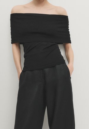 Person iført sort off-shoulder top og løse sorte bukser med hænderne i lommerne mod en ensfarvet lys baggrund.