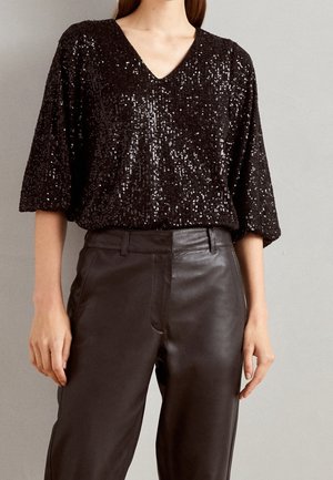 Sort paliet- V-hals top med albuelange ærmer kombineret med højtaljede mørke læderbukser på en stående person.