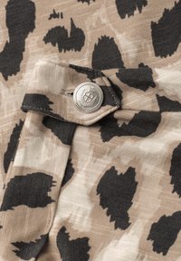 Gros plan sur un tissu beige avec des taches noires irrégulières et un bouton argenté cousu sur une patte de tissu.