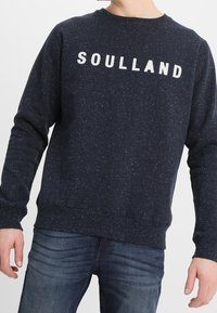 Felpa blu navy con logo bianco "SOULLAND", macchie testa di moro, scollatura rotonda e polsini e orlo a costine. Indossata sopra jeans blu.