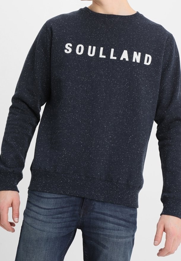 Felpa blu navy con logo bianco "SOULLAND", macchie testa di moro, scollatura rotonda e polsini e orlo a costine. Indossata sopra jeans blu.