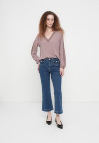 Μακρυμάνικο τοπ σε ανοιχτό μοβ χρώμα με V-λαιμόκοψη και δαντέλα, σε συνδυασμό με σκούρα μπλε τζιν καμπάνα. Το μοντέλο φορά μαύρα μυτερά παπούτσια.