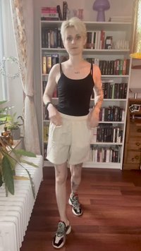 Persona con capelli biondi corti che indossa una canotta nera, pantaloni corti beige e sneakers, in piedi di fronte a una grande libreria in una stanza.
