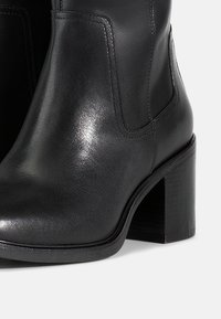 Botines de piel negra con una textura suave, punta redondeada y tacón cuadrado. Presentan costuras discretas y mínimos detalles de hardware.