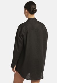 Schwarze Leinenbluse mit lockerer Passform, langen Ärmeln, geknöpften Manschetten und abgerundetem Saum. Schlichter Kragen und ein leichtes, strukturiertes Gewebe.