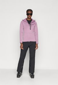 Chaqueta rosa con capucha, con cierre frontal, acentos negros y material de forro polar, combinada con pantalones de esquí negros y botas de esquí.