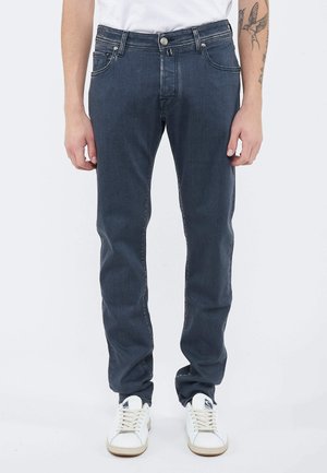 Uomo che indossa jeans blu scuro, sneakers bianche e una camicia bianca, in piedi con le braccia rilassate lungo i fianchi, tatuaggio visibile sull'avambraccio sinistro.