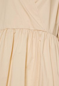 Abito beige con gonna arricciata. Presenta un tessuto liscio e leggero e un design avvolgente, con cuciture visibili lungo le giunture.