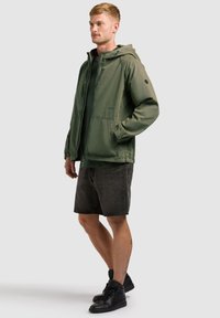 Homme debout portant une veste à capuche verte, un short en denim gris foncé et des baskets montantes noires sur un fond uni.