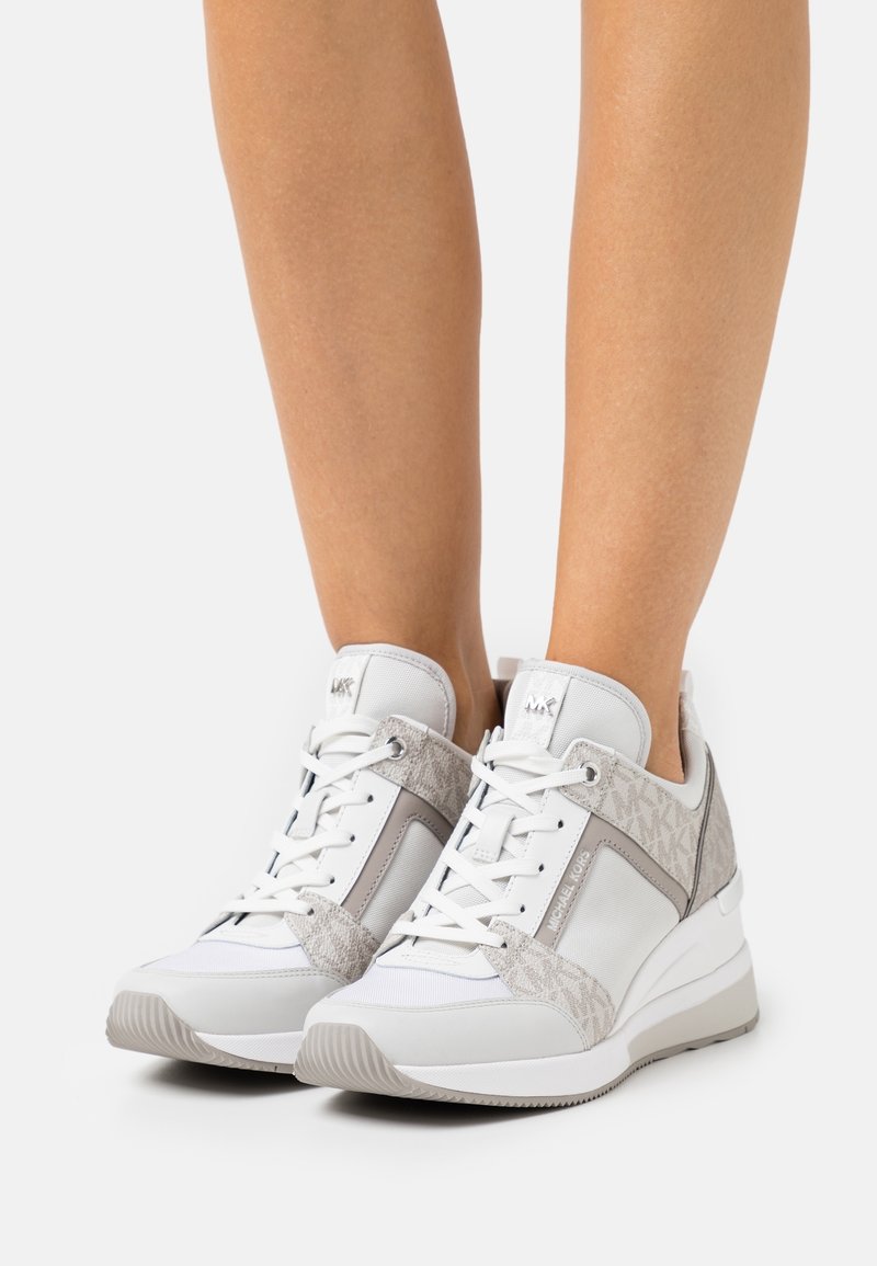 Personne portant des baskets compensées blanches et beige clair avec détails du logo "MK", lacets noués, debout sur fond blanc.