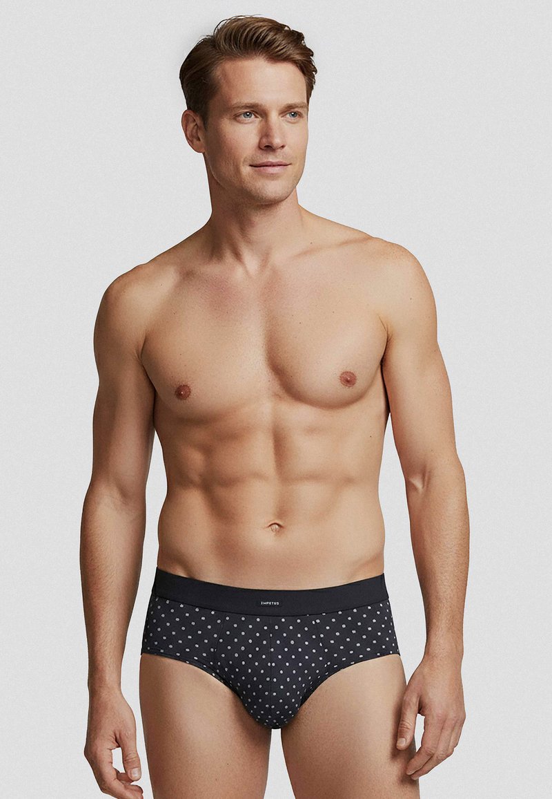 Uomo muscoloso che indossa slip neri con un piccolo motivo a pois bianco, guardando leggermente a destra su uno sfondo chiaro uniforme.