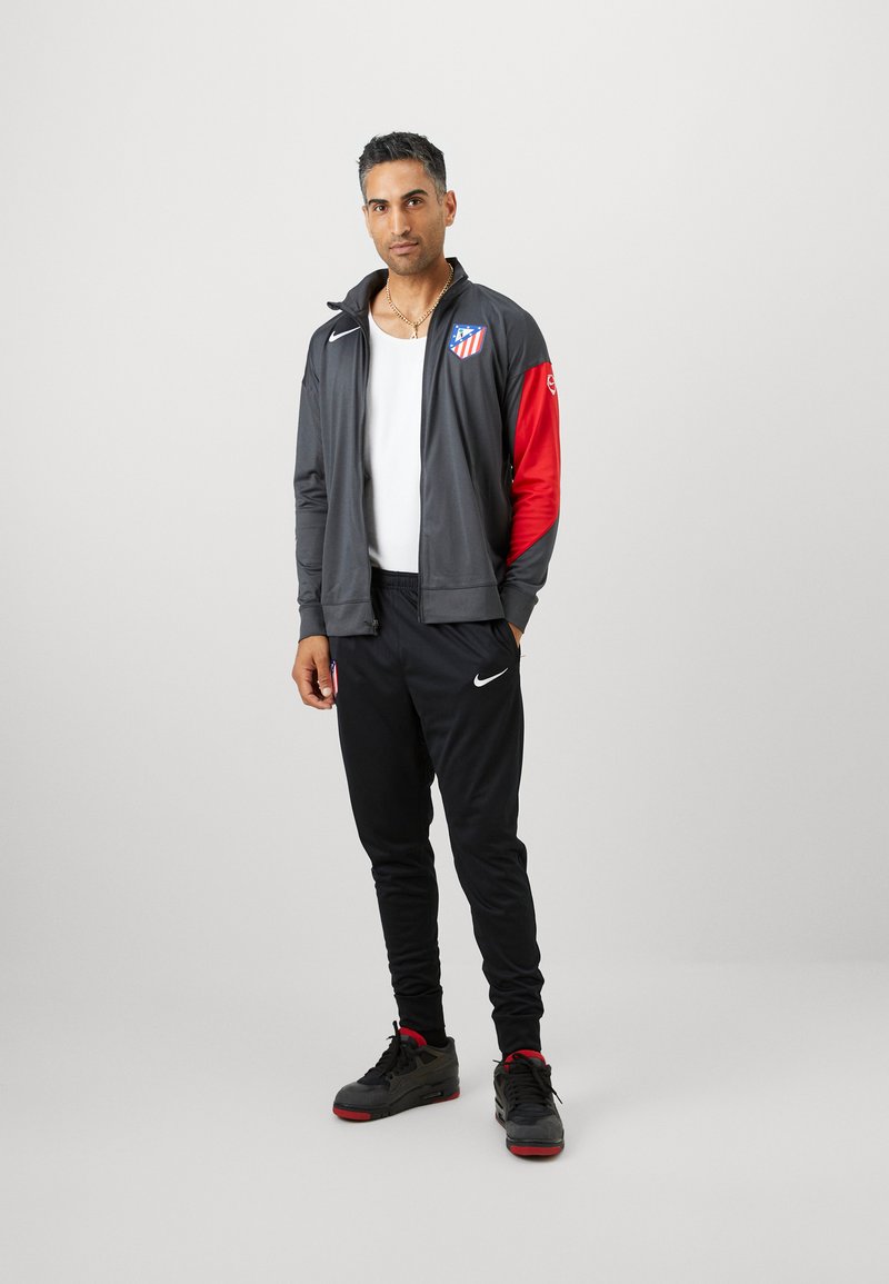 Veste de sport grise avec des manches rouges, arborant un logo. Associée à un pantalon noir et des chaussures noires avec des accents rouges, le tout dans un design ajusté.