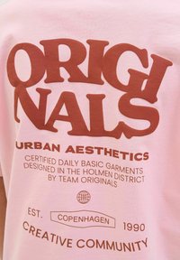 Rozā kokvilnas T-krekls ar lielu brūnu tekstu, kas saka "ORIGINALS URBAN AESTHETICS", un papildu tekstu un globusa grafiku.