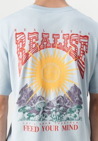 Lichtblauw katoenen t-shirt met een grafisch ontwerp van een gele zon, bergen en paddenstoelen; rode blokletters bevatten de tekst "REALISE" en "FEED YOUR MIND."