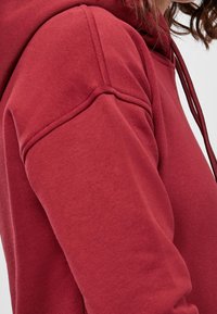 Sudadera de algodón roja con capucha ajustable, puños acanalados y una textura suave con costuras visibles a lo largo de la costura del hombro.