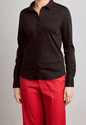 Osoba nosząca dopasowaną czarną koszulę zapinaną na guziki z długim rękawem i jaskrawo czerwone spodnie stojąca na jednolitym jasnoszarym tle.