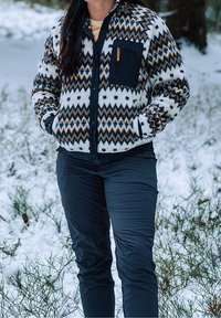 Personne portant une veste polaire à motifs et un pantalon foncé, debout les mains dans les poches dans un environnement extérieur enneigé.